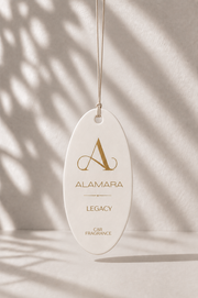 Oval-shaped 'Alamara Legacy' car fragrance hanging against a neutral background with leaf shadows.